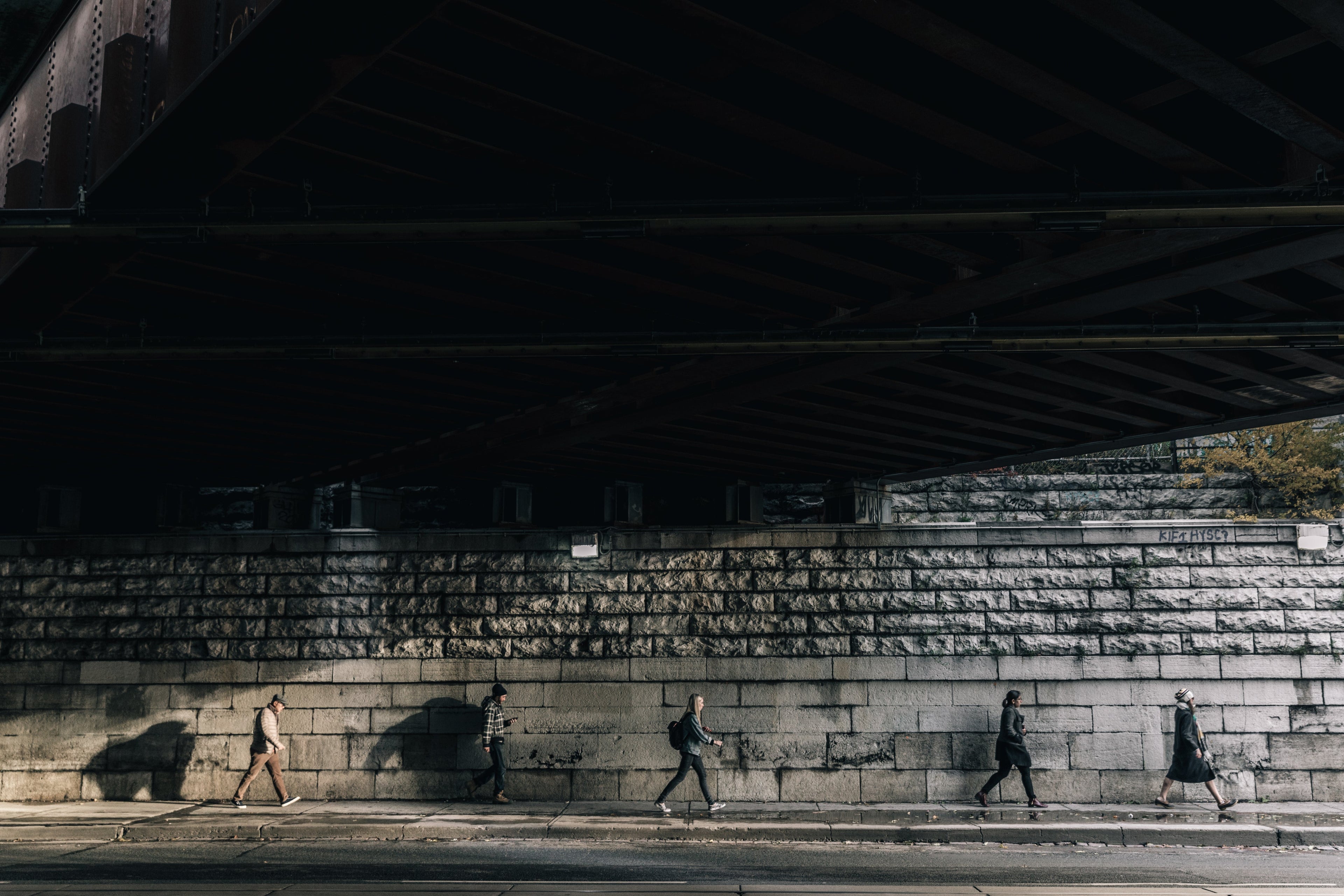 People walking under a bridge with a stone wall in the background