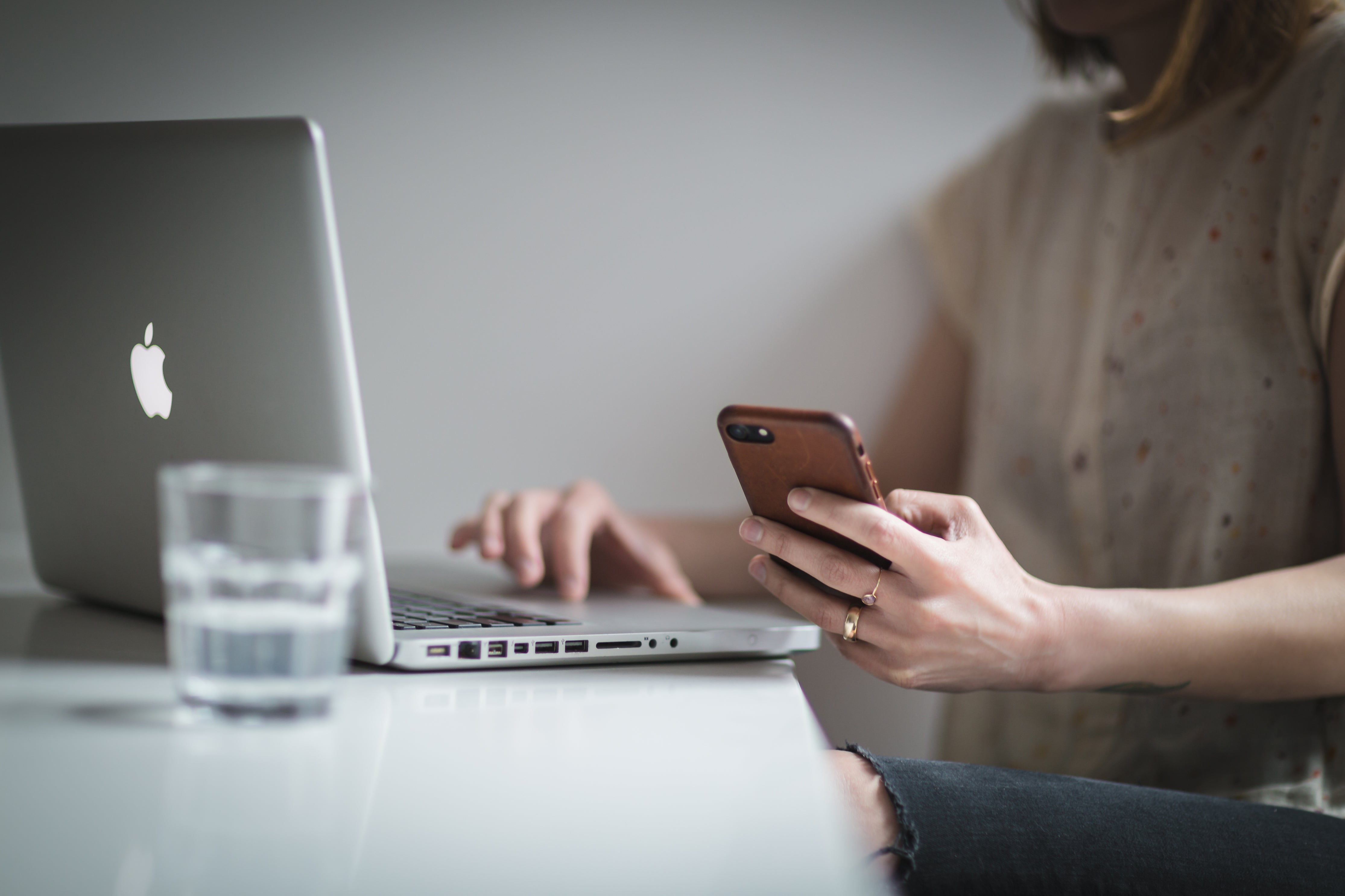 Person using a laptop and smartphone at a desk with a glass of water.